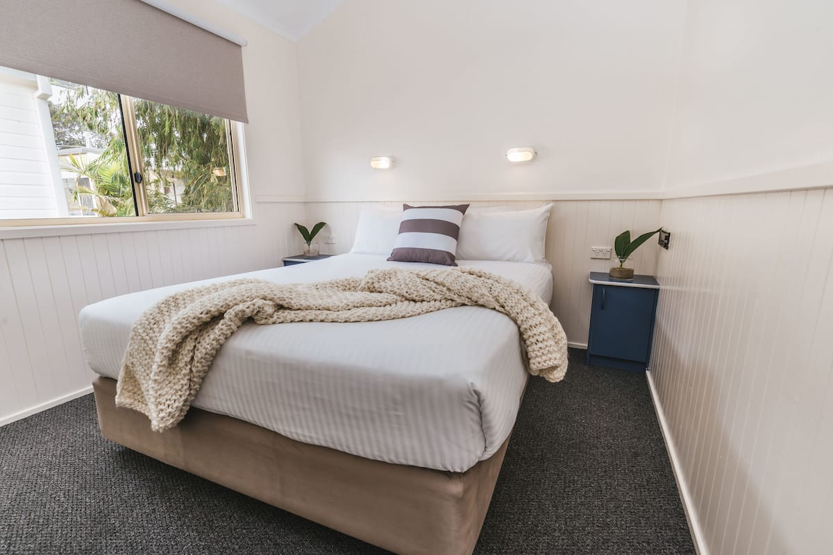 Cama bem arrumada com um cobertor bege, duas mesas de cabeceira com plantas e uma janela com persiana.