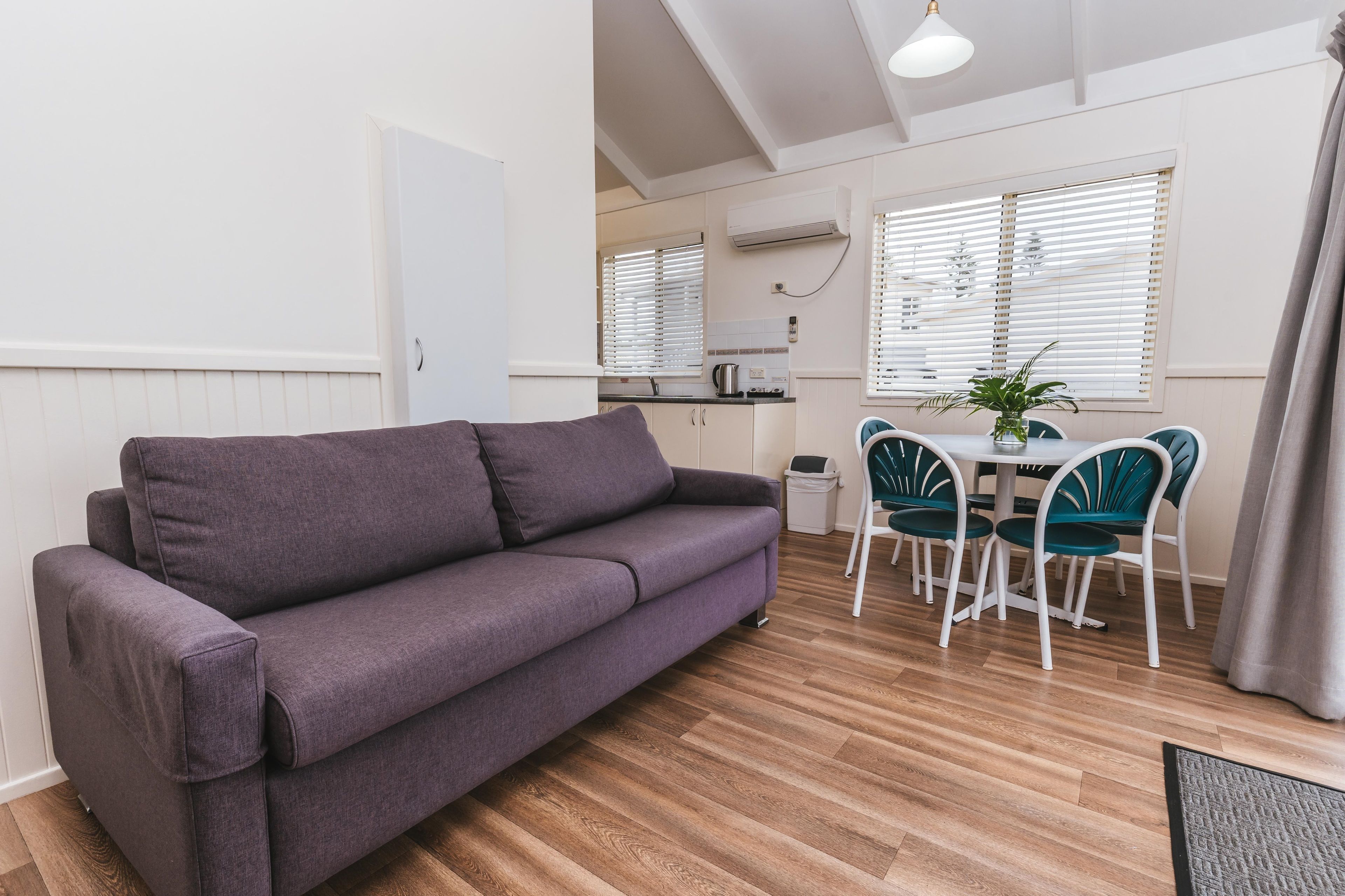 A modern living room with a grey sofa, a dining area with a white table and teal chairs, and wooden flooring.