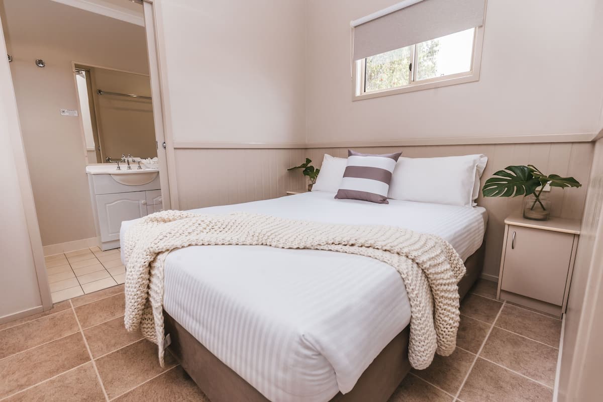 Quarto com cama grande, mesa de cabeceira, janela e banheiro visível ao fundo.