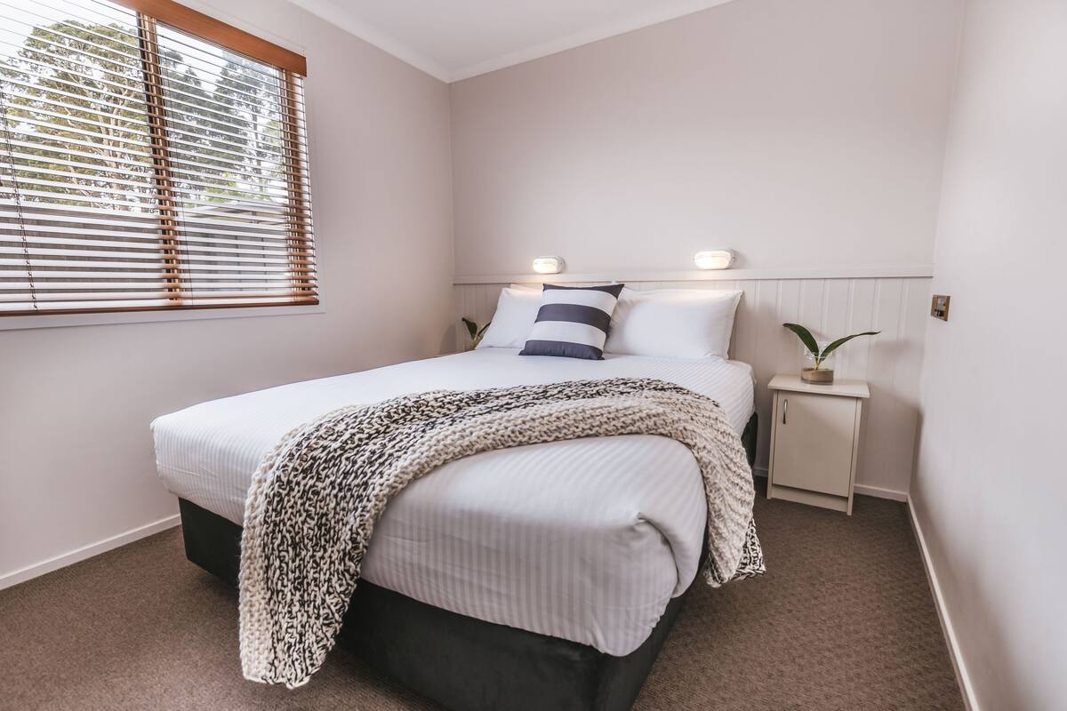 Quarto com cama, mesas de cabeceira e janela com persianas.