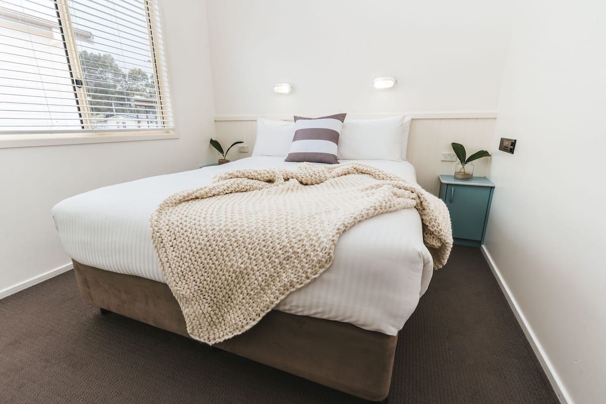 Cama bem arrumada com manta bege, duas mesas de cabeceira com plantas e uma janela com persianas.