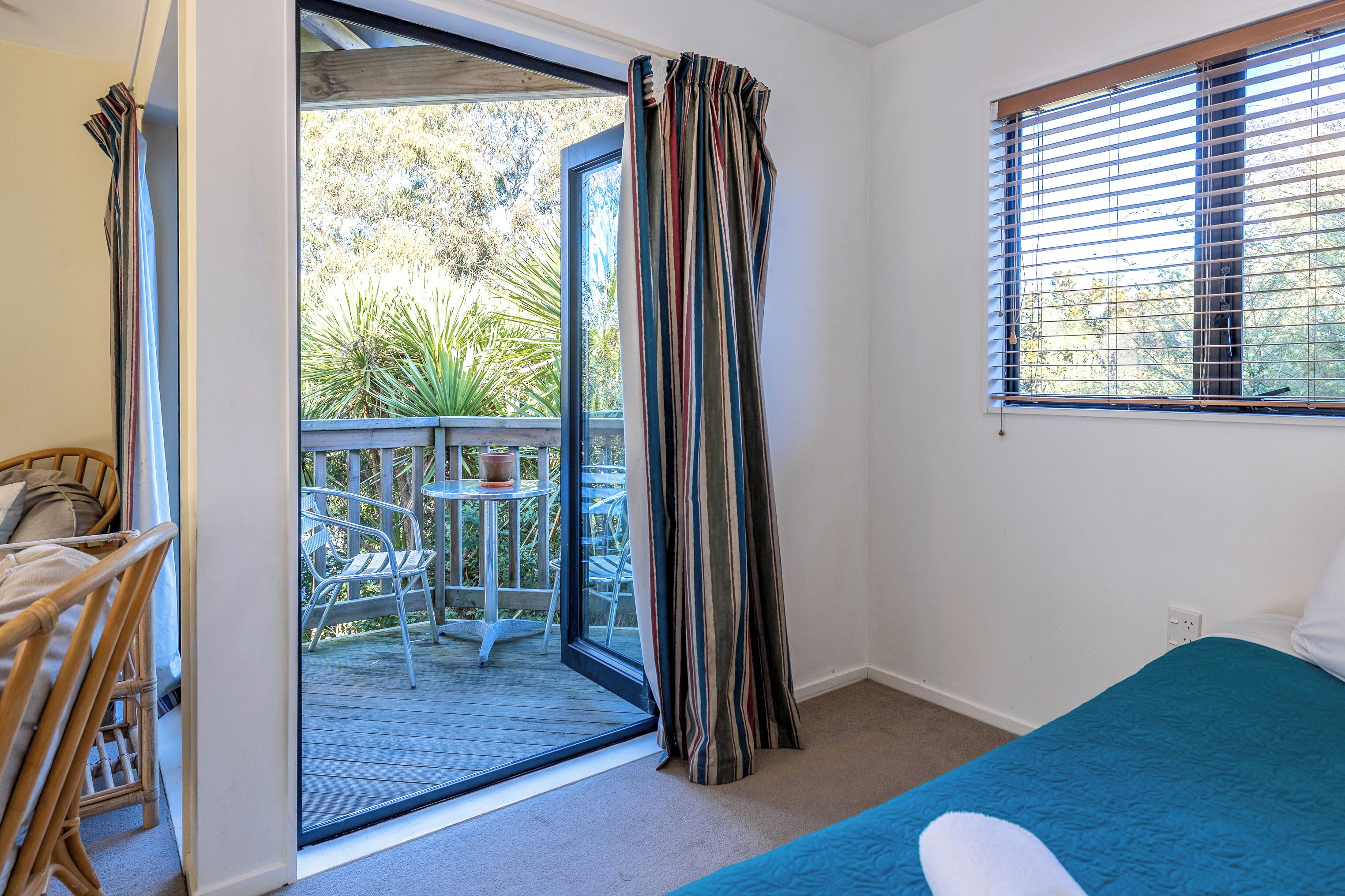 A room with a bed, a window with blinds, a sliding glass door leading to an outdoor area with a table and chairs, and a view of greenery outside.
