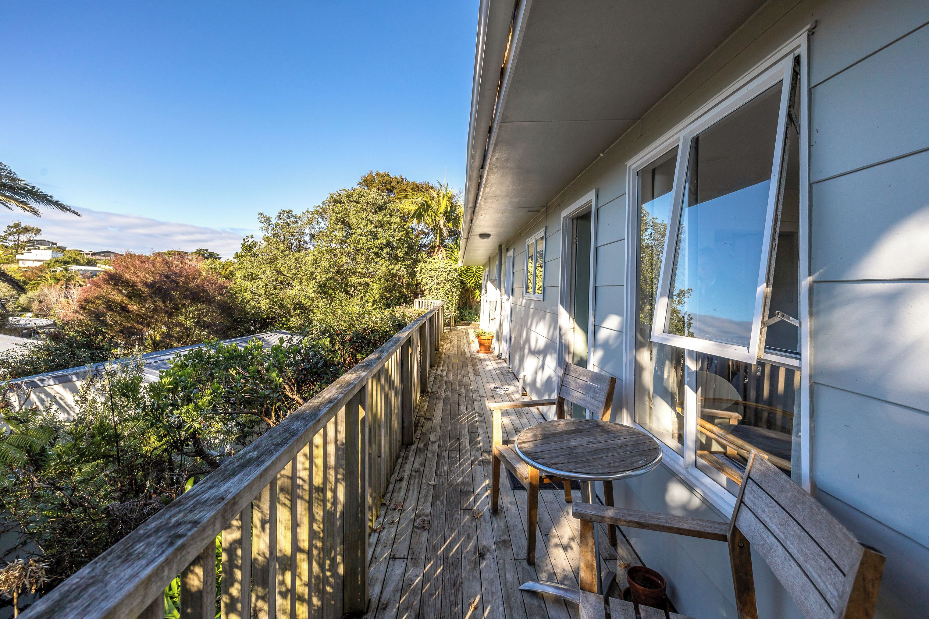 Una terraza de madera con una mesa y sillas, con vistas a un jardín exuberante y una carretera.