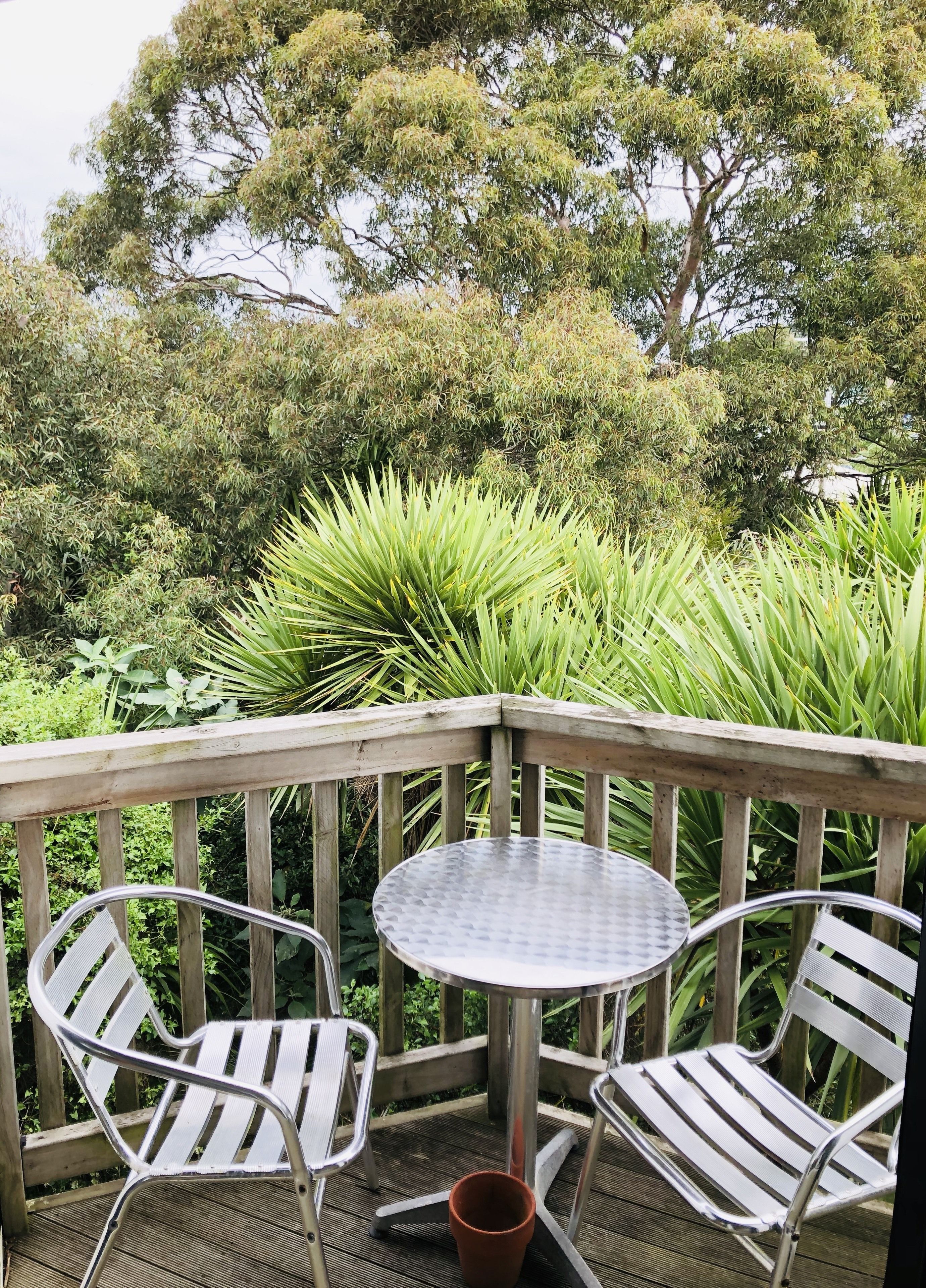 A wooden deck with two metal chairs and a small table, surrounded by lush greenery and trees.
