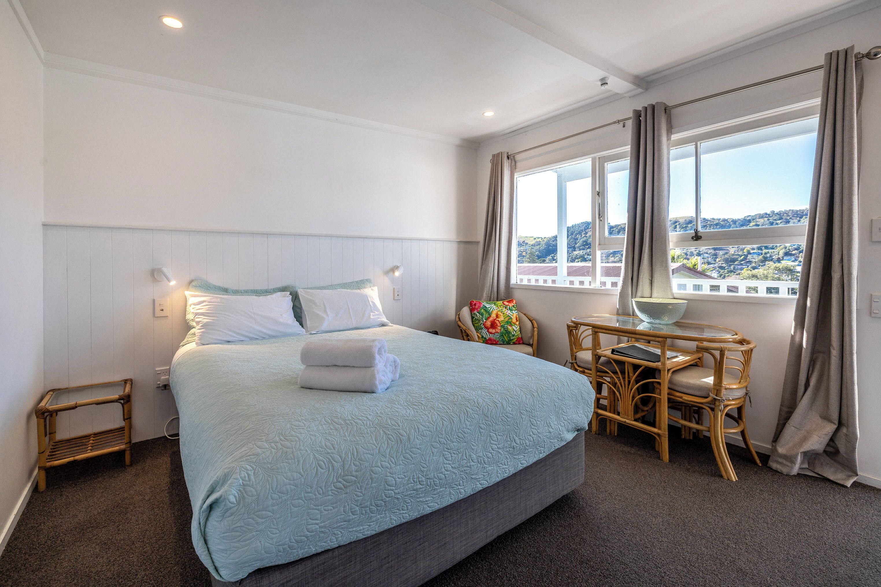 A hotel room with a large bed, a small table with chairs, a window with curtains, and a view of a cityscape.