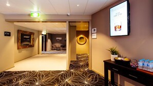 A hotel lobby with a patterned carpet, a reception desk with a plant and water bottles, an ATM sign, and a digital screen displaying a welcome message. - Shamrock Gardens Motel (West Mackay)