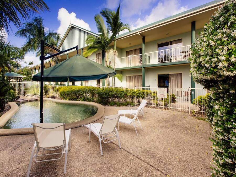 A pool area with lounge chairs and umbrellas, surrounded by a building with balconies and palm trees.
