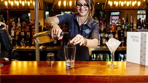 A bartender pouring a drink into a glass at a bar with a drinks menu sign in the background. - Shamrock Gardens Motel (West Mackay)