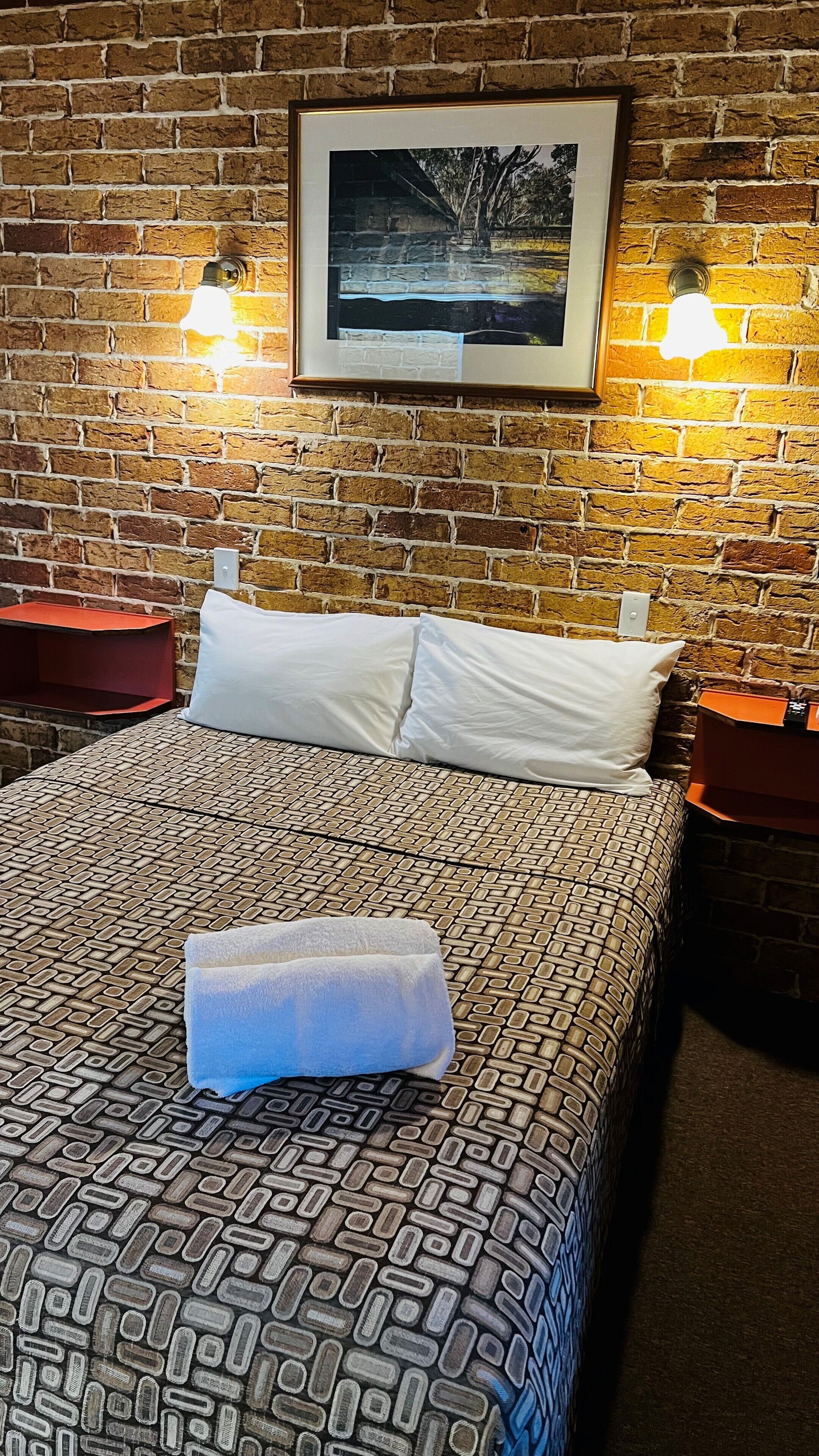 A hotel room with a brick-patterned bedspread, two white pillows, a framed picture on the wall, and two nightstands with red lamps.