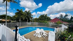 A swimming pool with a white fence, surrounded by palm trees and a red-roofed building. - Beenleigh Village Motel (Beenleigh)
