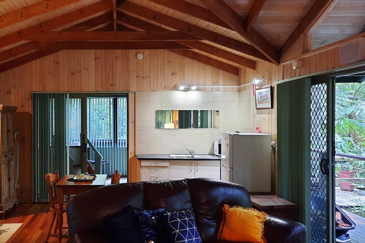 A wooden cabin interior with a dining area, kitchen, and a large window overlooking greenery.