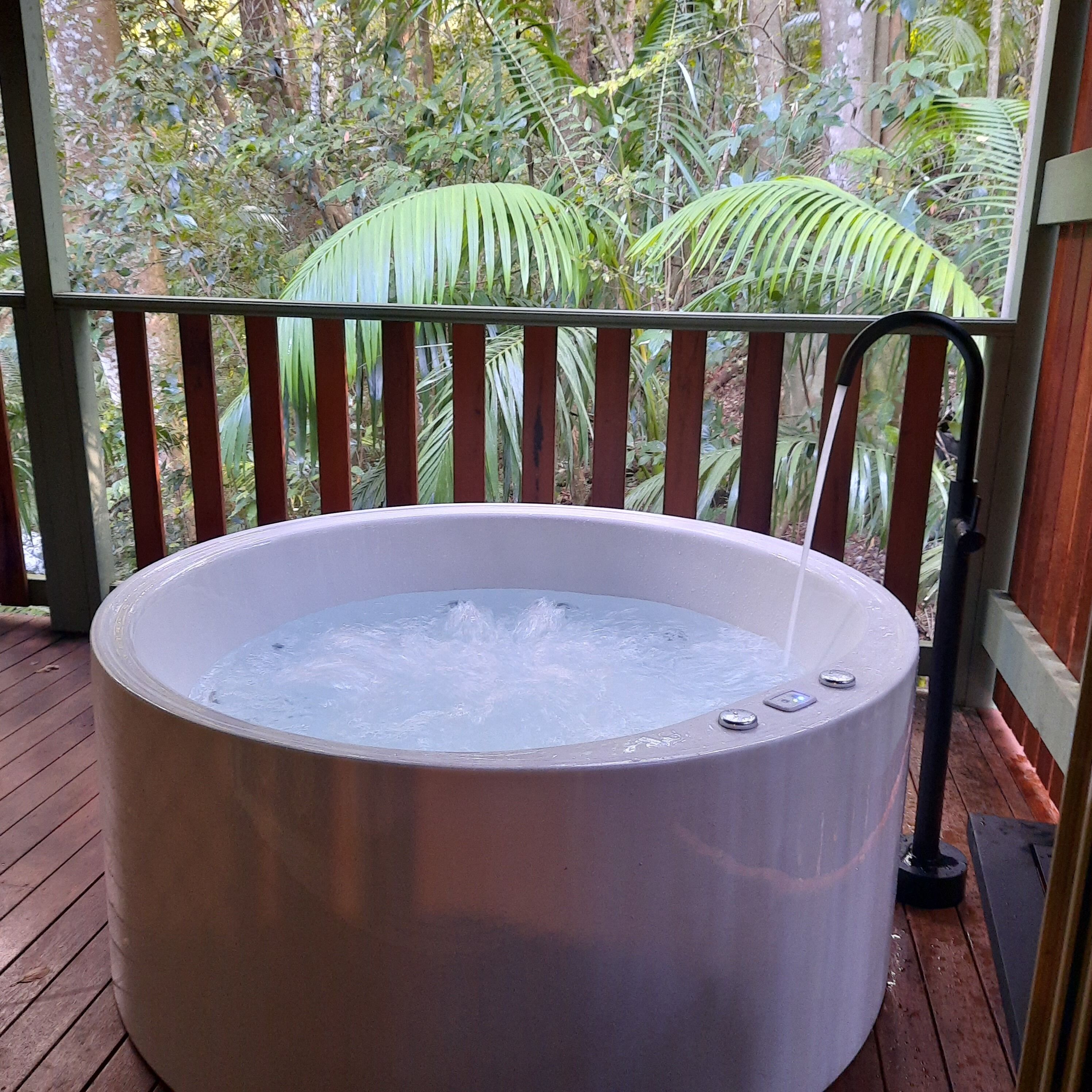 Ein Whirlpool auf einer Holzterrasse mit Geländer, umgeben von üppigem Grün.
