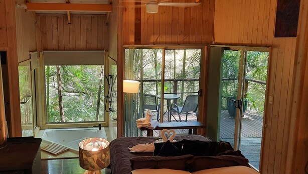 A wooden cabin with a large glass door leading to an outdoor deck. Inside, there is a bed with a heart-shaped pillow and a table with a lamp. - Treetops Accommodation Montville (Flaxton)