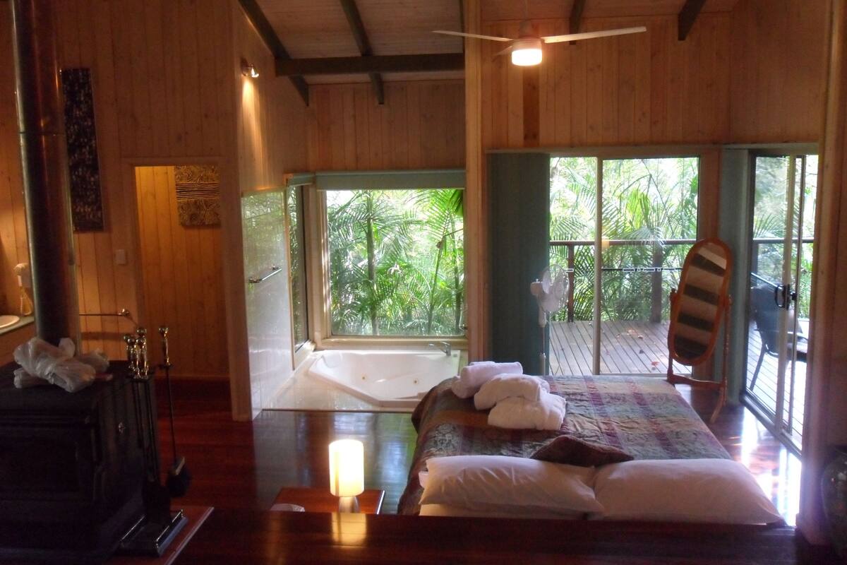 A wooden room with a bed, a bathtub, and a view of greenery outside.