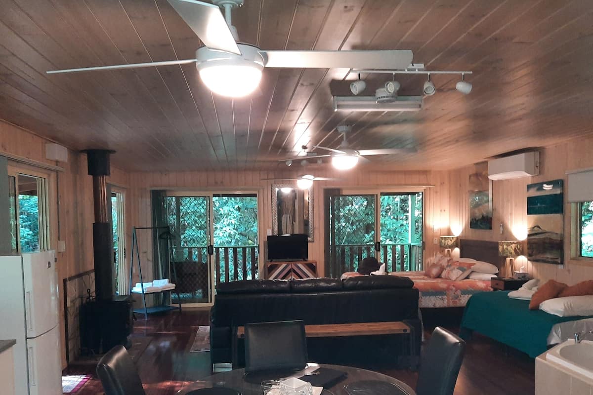 A spacious room with a dining table, chairs, a sofa, and a bed. There is a ceiling fan, a window with a view of trees, and a fireplace.
