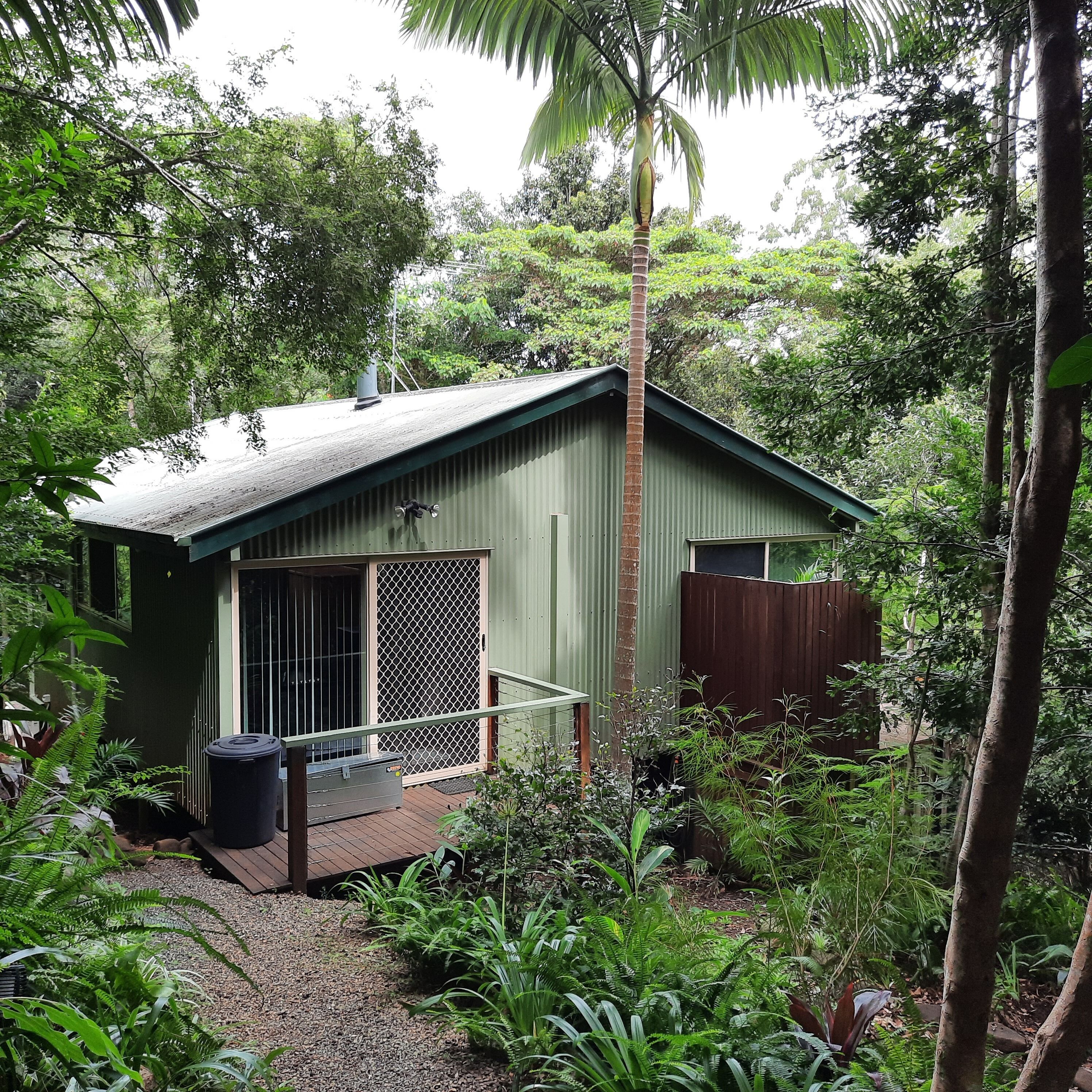 A small, single-story building with a green roof and a metal fence, surrounded by dense greenery and trees.
