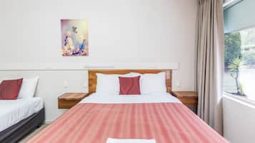 A hotel room with two beds, a large window with curtains, a painting on the wall, and a bedside table with a lamp.