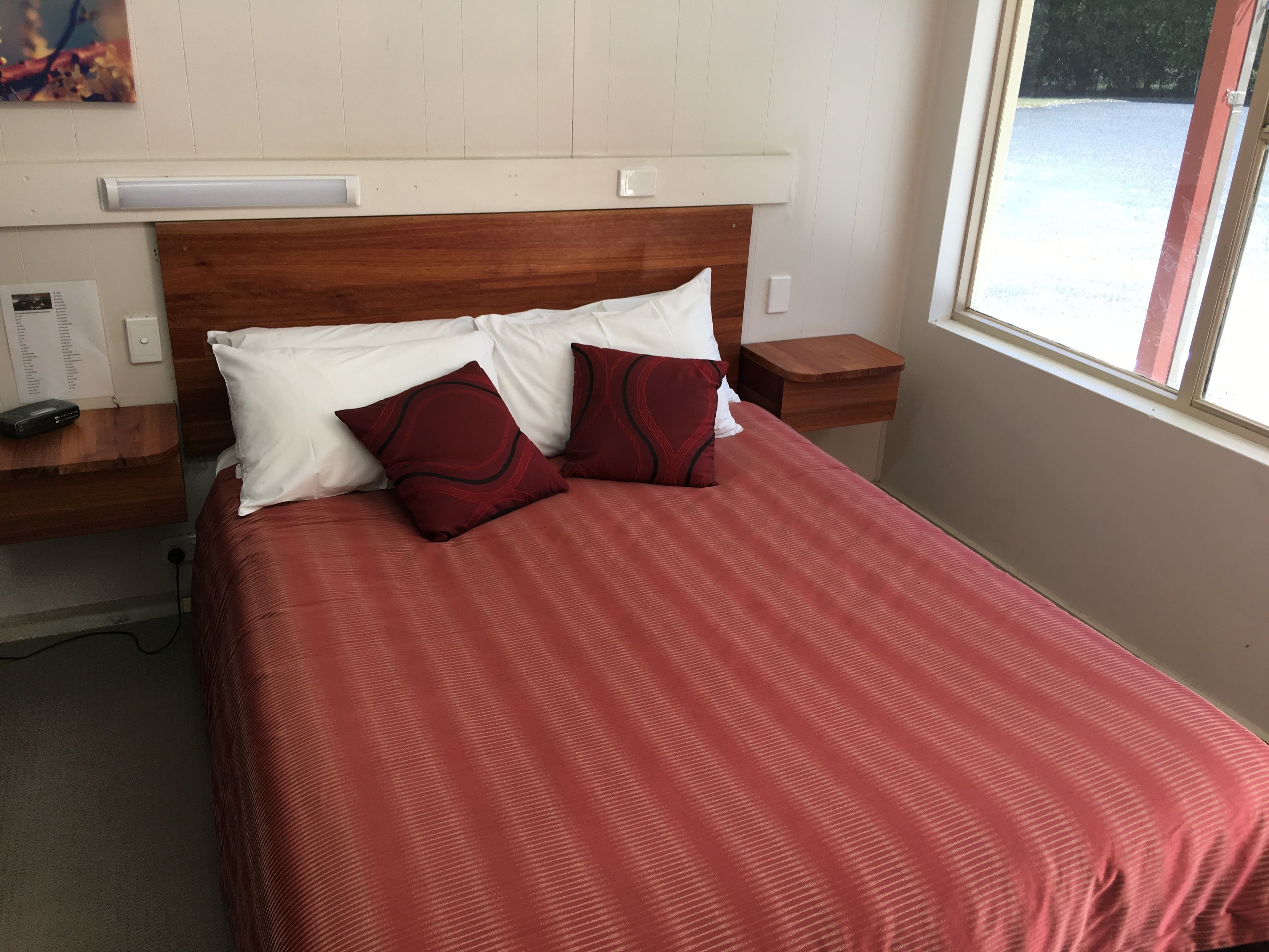 Una cama bien hecha con una colcha roja y dos almohadas, un cabecero de madera, una mesita de noche con una lámpara y una ventana con vistas a un paisaje nevado.
