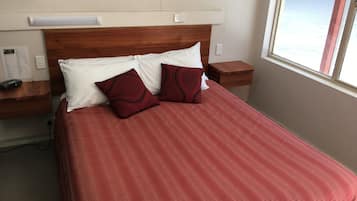 Una cama bien hecha con una colcha roja y dos almohadas, un cabecero de madera, una mesita de noche con una lámpara y una ventana con vistas a un paisaje nevado.
