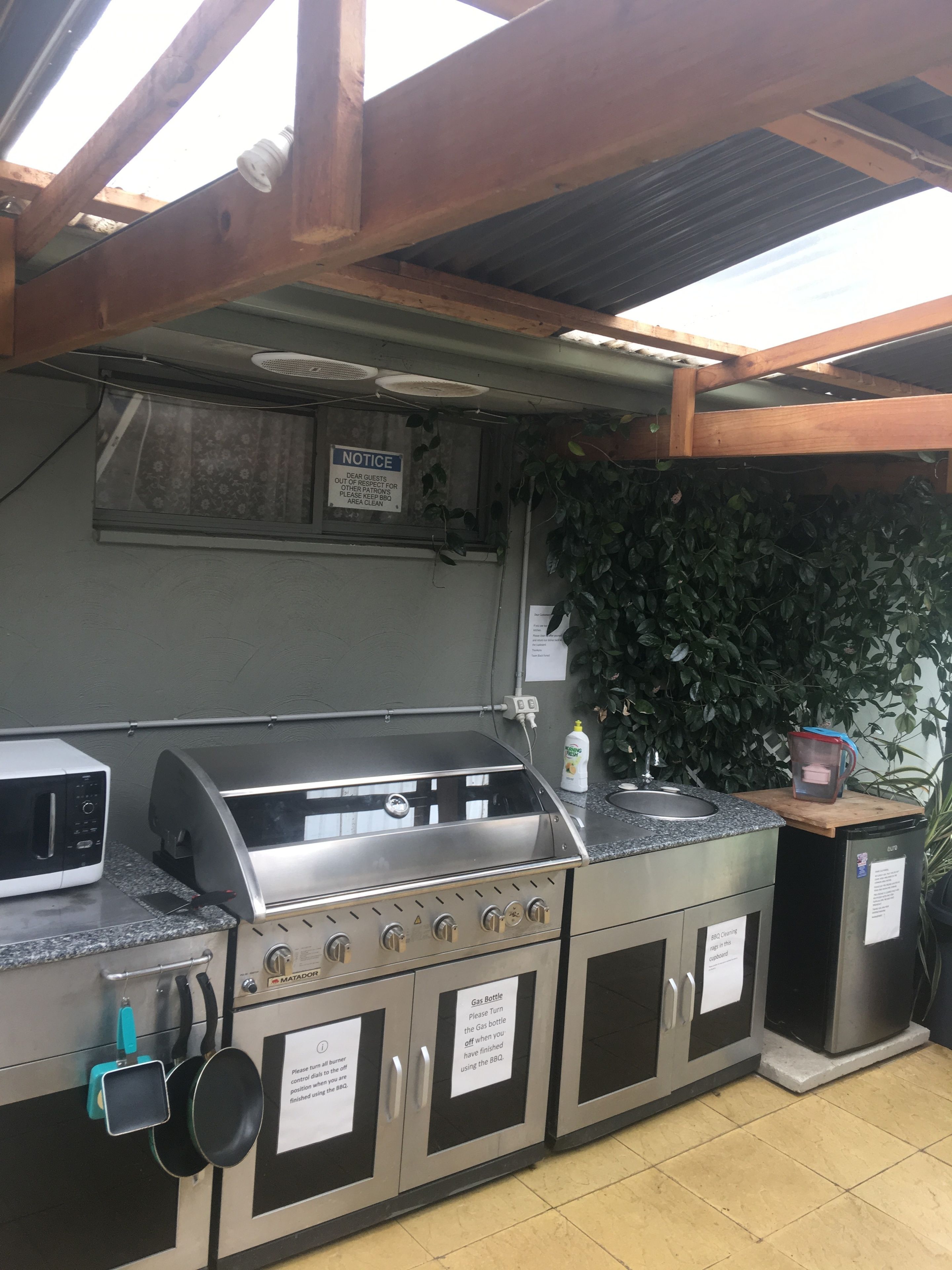 Cocina exterior con parrilla a gas, microondas y pileta. Hay un aviso en la pared y un cartel que indica el área de barbacoa.