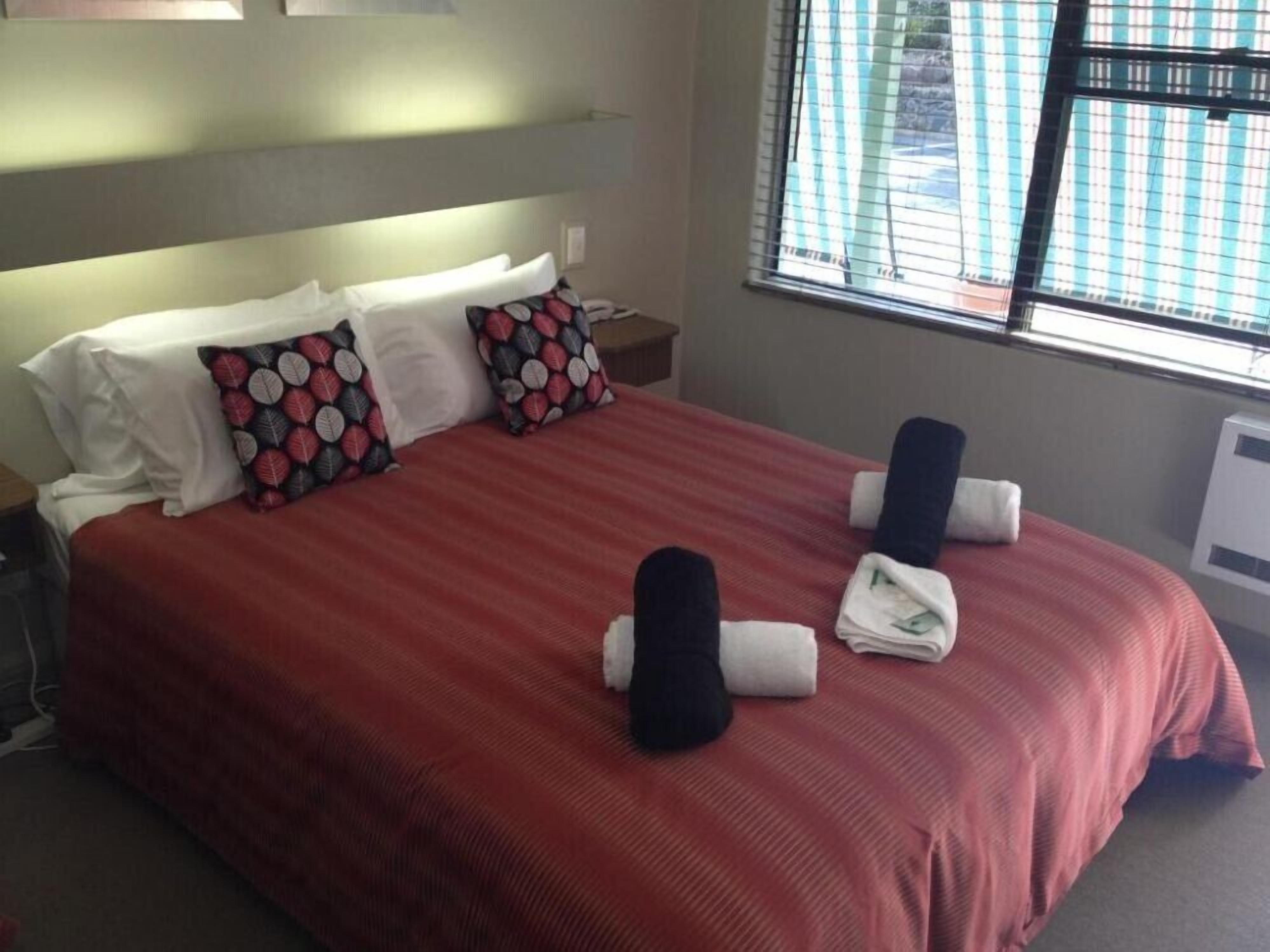 A hotel room with a large bed, two pillows, a window with blinds, and a bedside table.