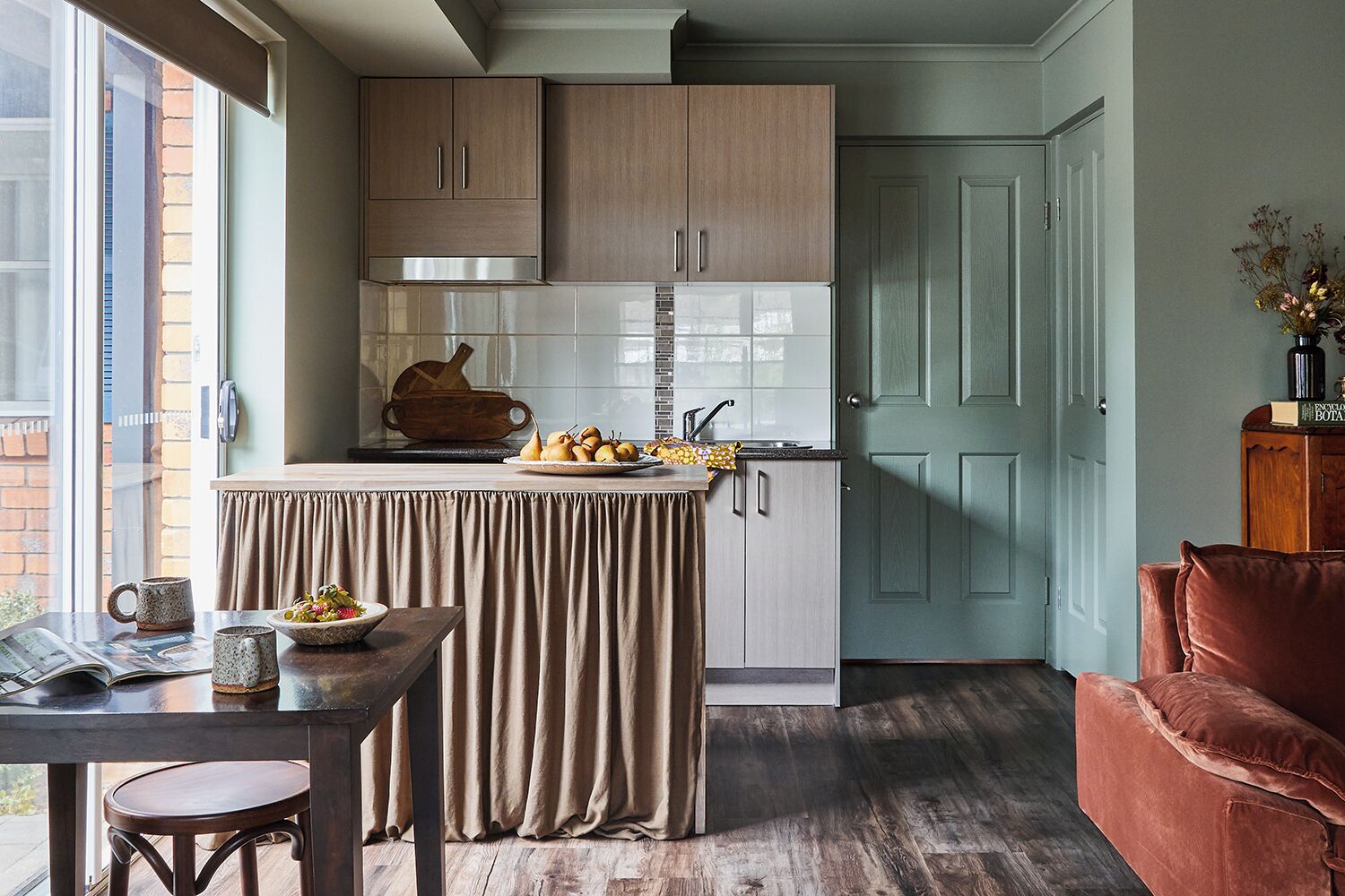 Una cucina compatta con isola centrale, zona pranzo con tavolo e sedie, e angolo relax con poltrona imbottita.