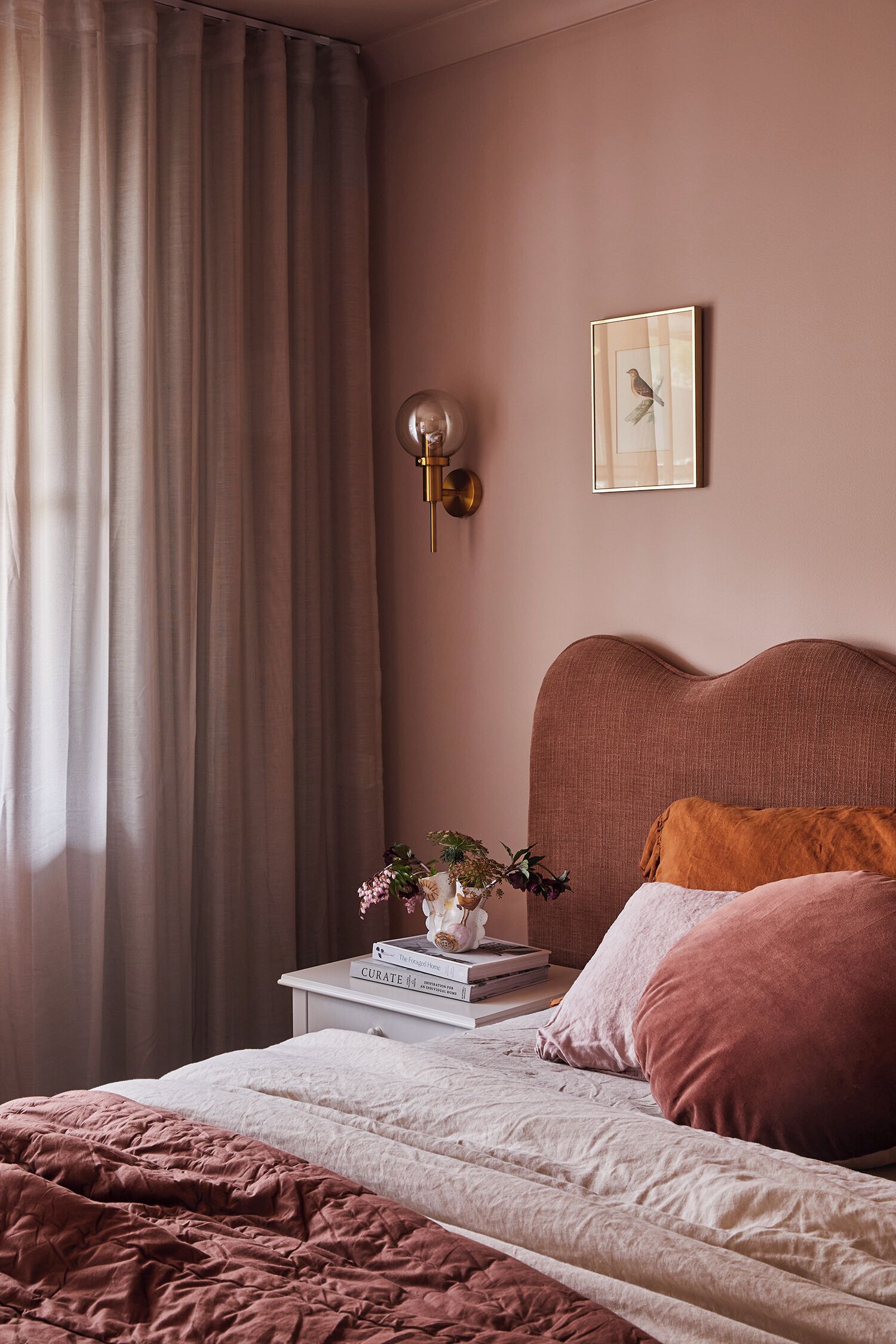 A bedroom with a bed, a nightstand with books and a vase, a wall-mounted light, and a framed picture.
