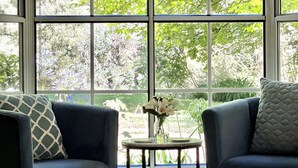 A room with two blue armchairs, a small table with a vase of flowers, and a large window with a view of green trees.