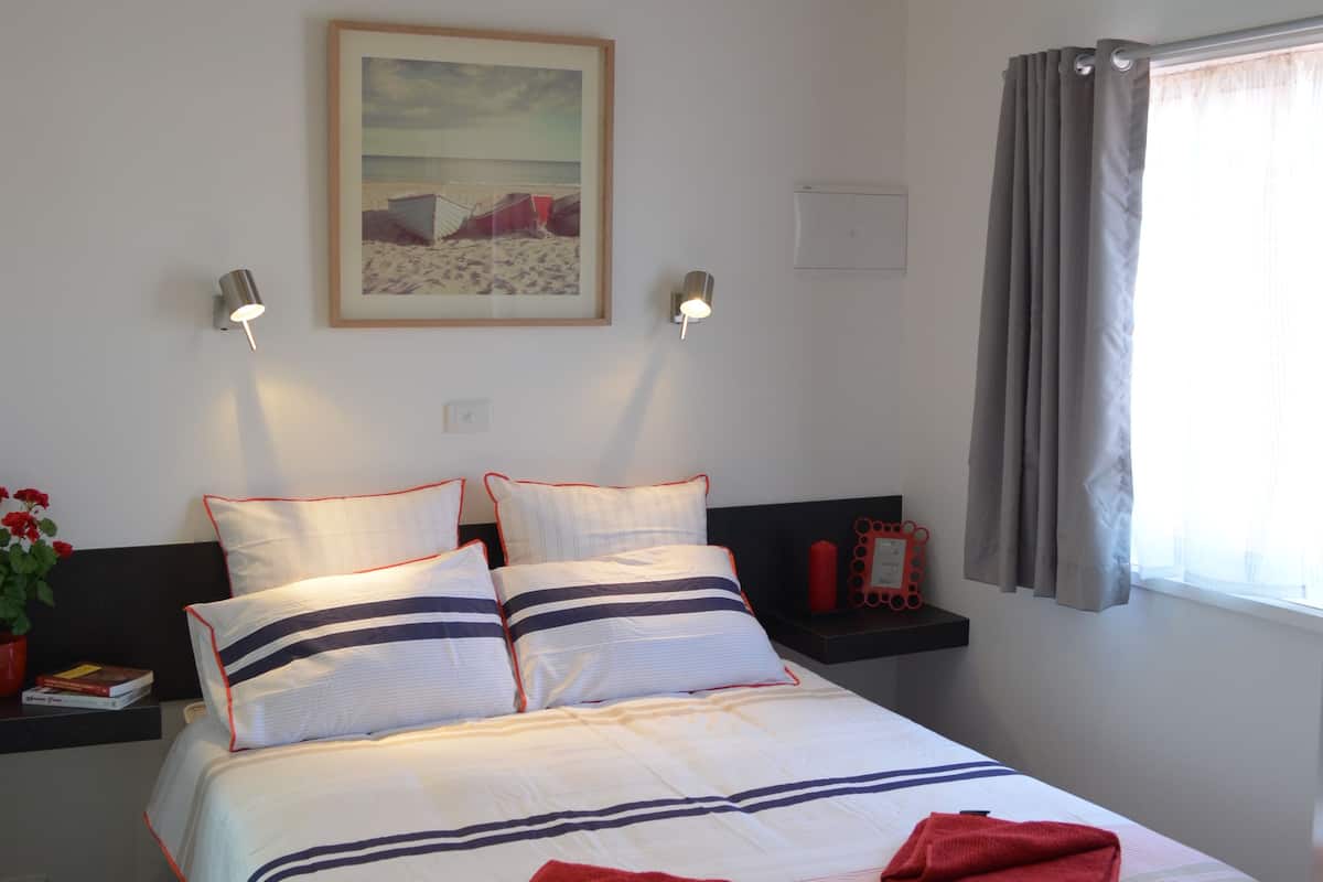 A neatly made bed with a striped bedspread, pillows, and two red towels. There is a ceiling fan, a wall-mounted light, and a framed picture above the bed.