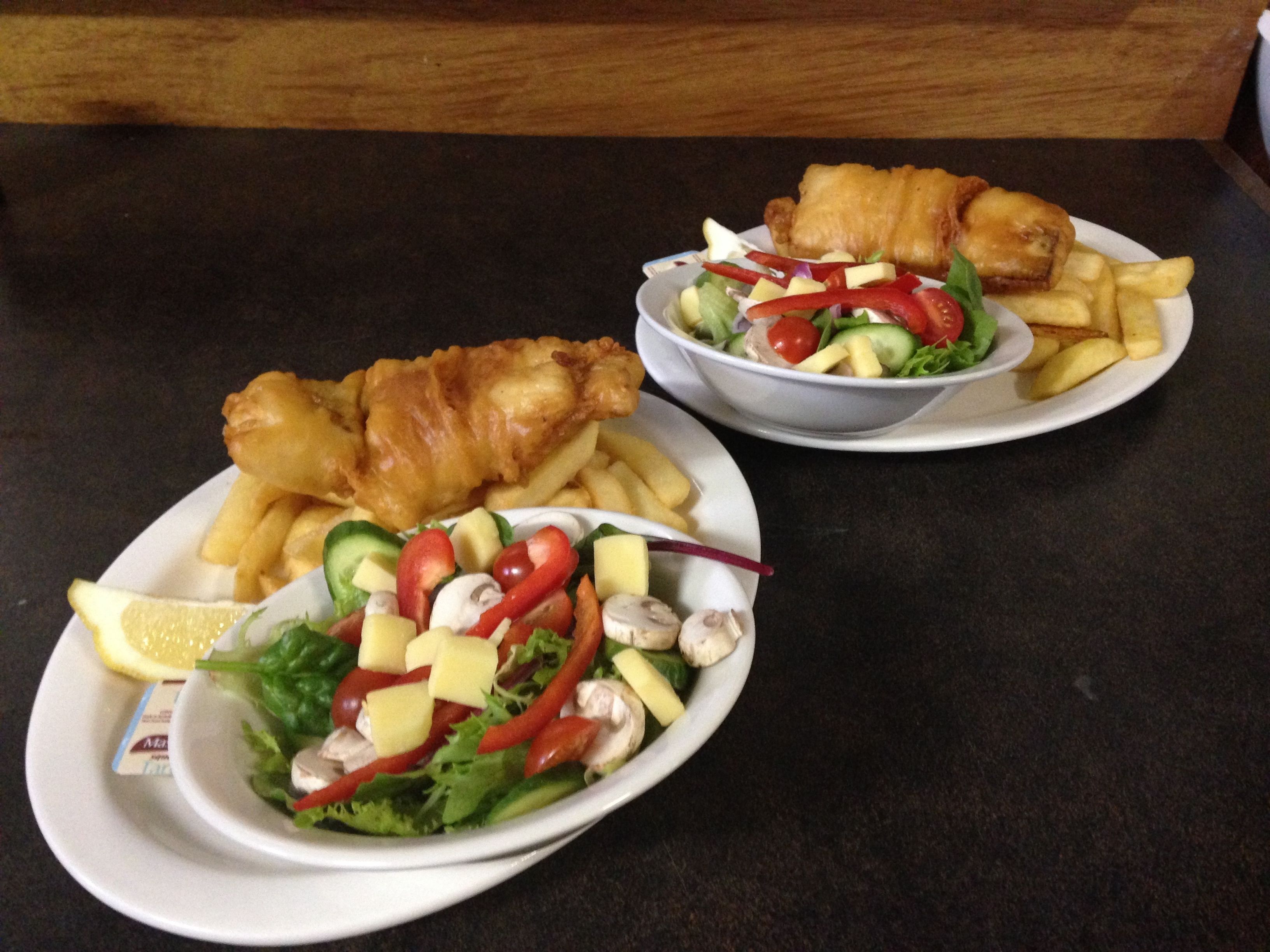 Una mesa con dos platos de comida, uno con pescado y papas fritas y una ensalada, y otro con una comida parecida.