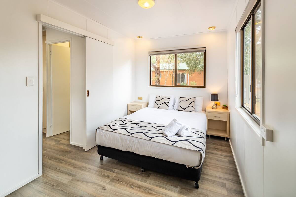 Un dormitorio con una cama, mesitas de noche, una ventana y una puerta.
