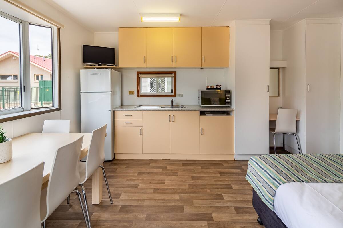 Cocina compacta con refrigerador, microondas y pileta. Cuenta con una mesa de comedor con sillas y una cama con cubrecama a rayas.