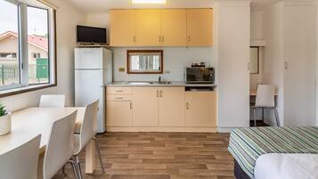 A compact kitchen with a refrigerator, microwave, and sink. There is a dining table with chairs and a bed with a striped bedspread.