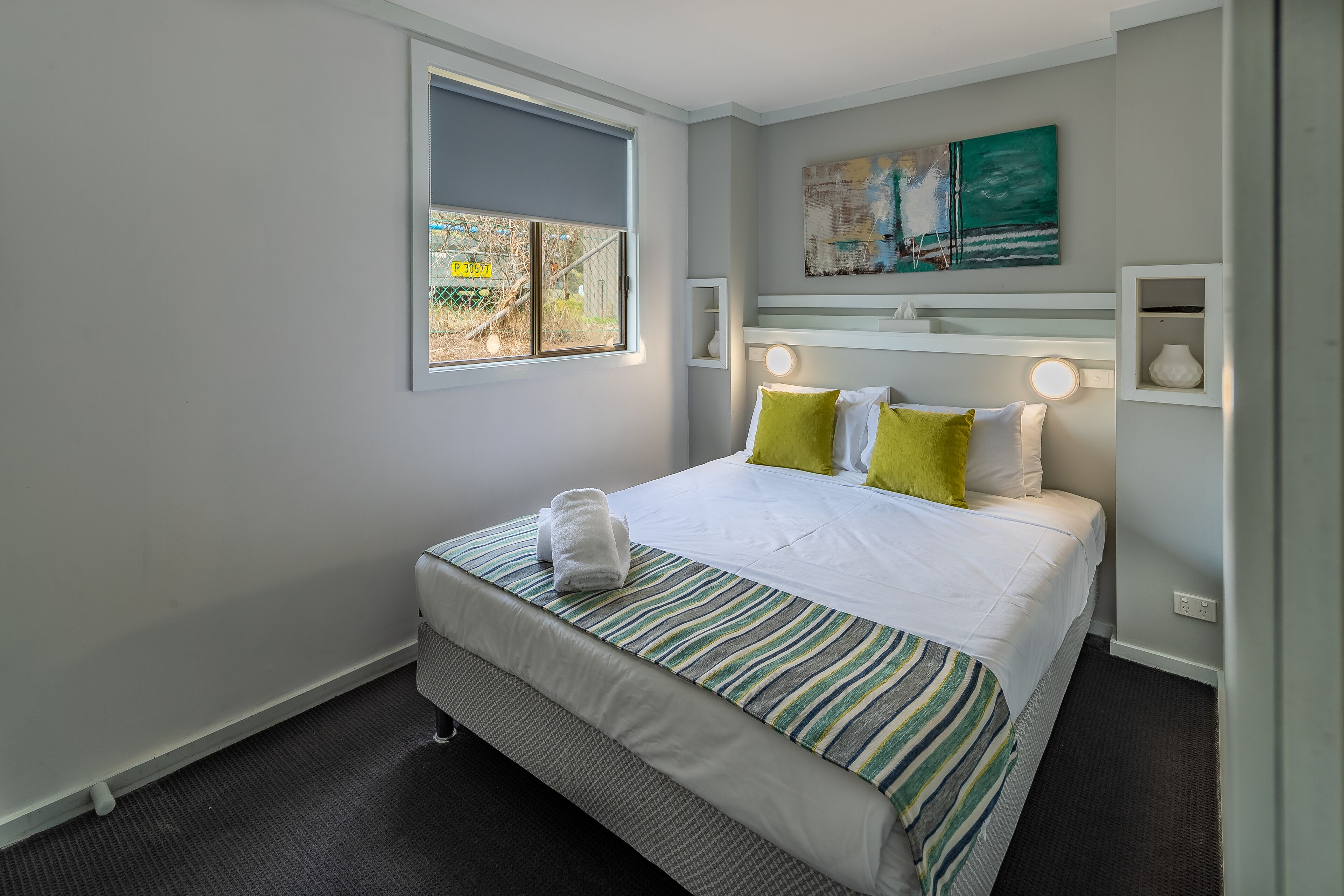 A neatly arranged hotel room with a bed, a window, a painting, and a shelf with decorative items.