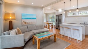 A modern living room with a sofa, a coffee table, and a painting of a beach scene. There is a kitchen area with a bar and stools. - NRMA Broulee Holiday Park (Broulee)