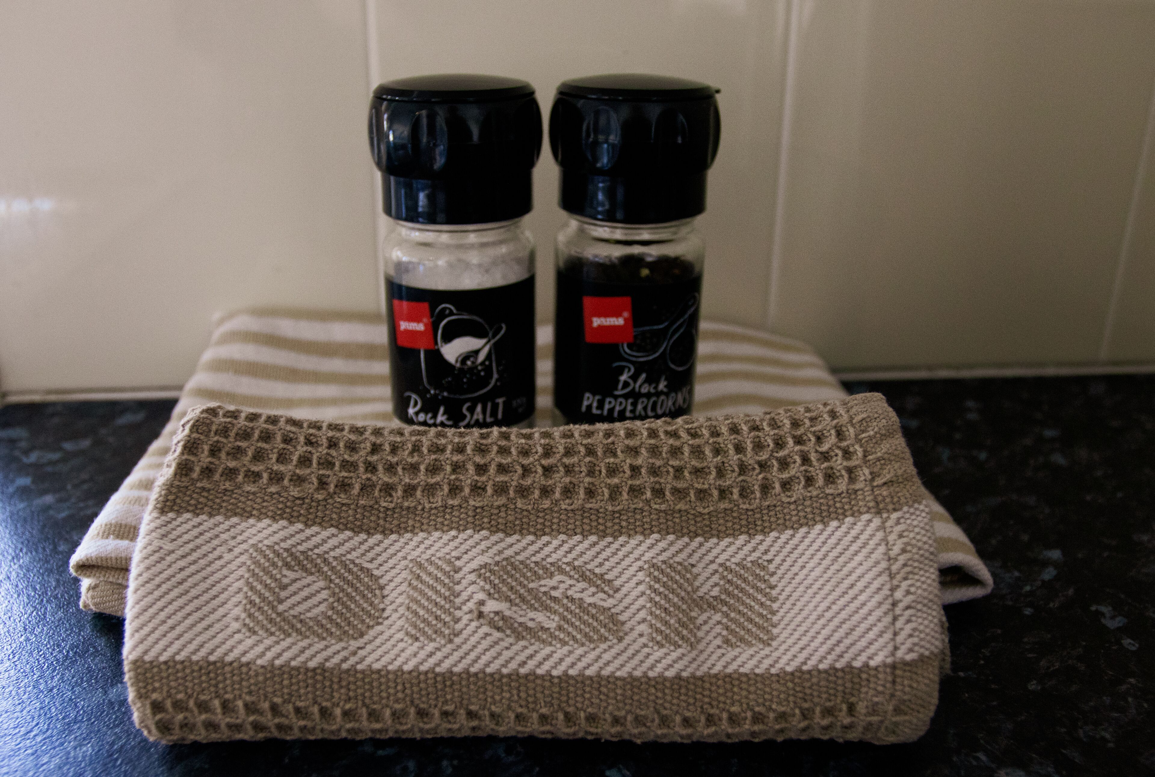 A kitchen counter with a woven placemat, two spice jars labeled 'Rock Salt' and 'Black Peppercorns', and a striped cloth.