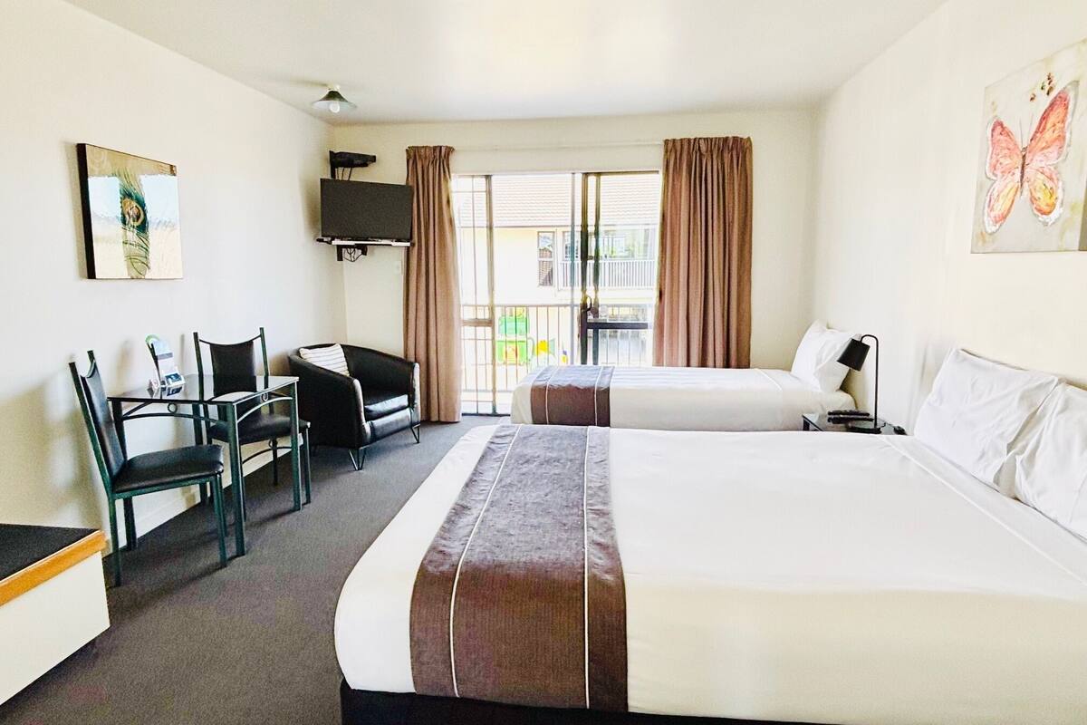 A hotel room with two beds, a desk with a chair, a TV mounted on the wall, and a balcony with curtains.