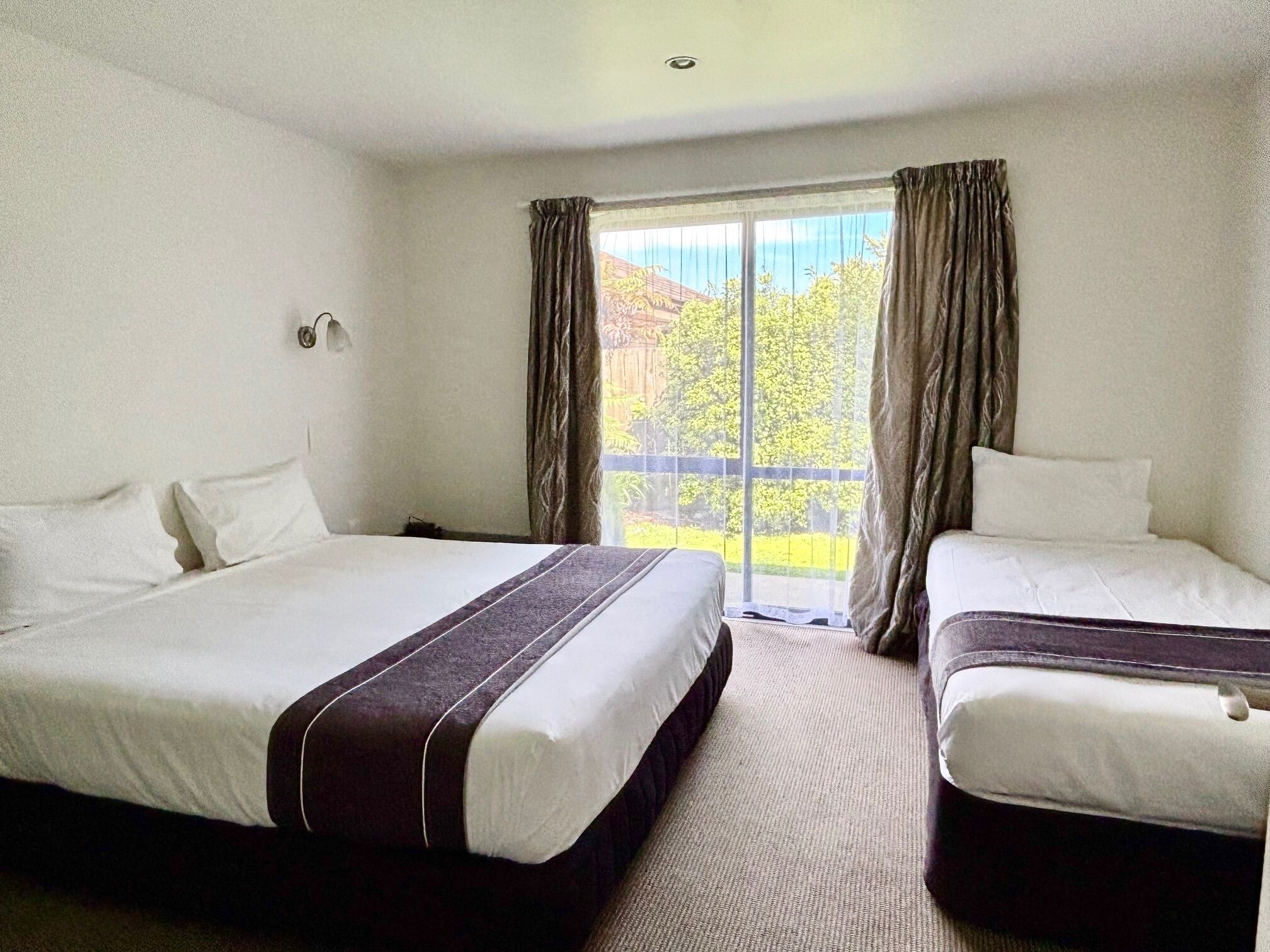 A hotel room with two beds, a large window with curtains, and a view of greenery outside.