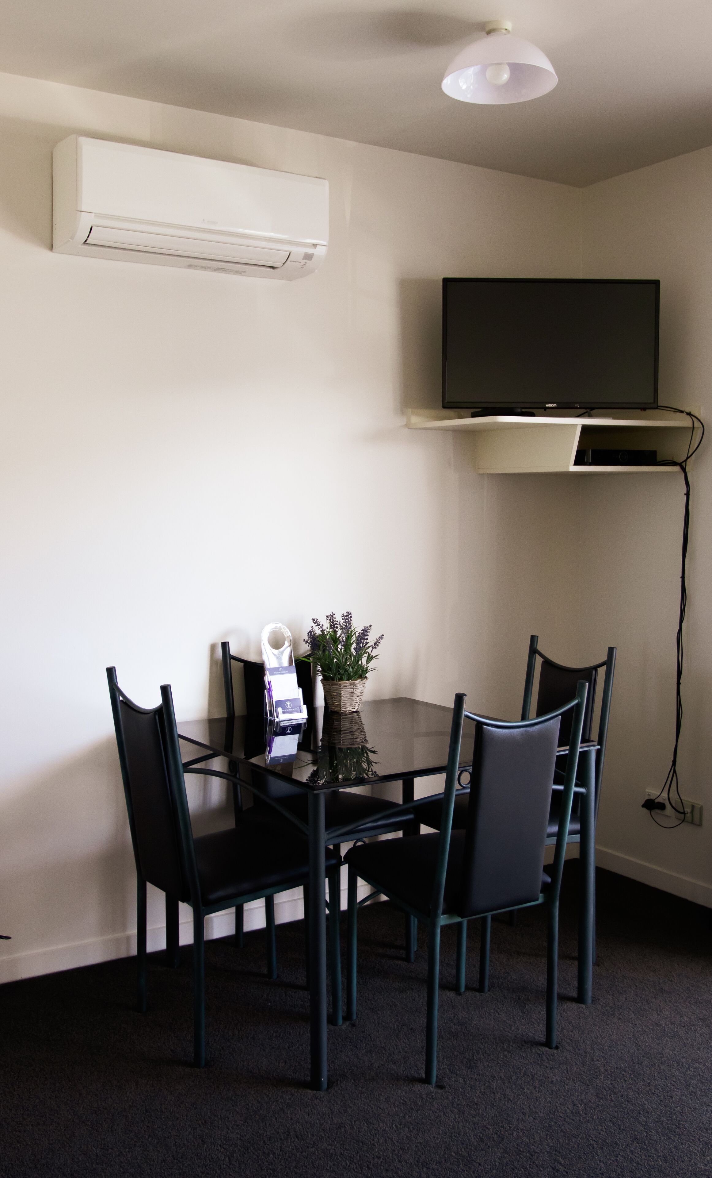 A small dining area with a glass table, four chairs, a potted plant, and a mounted flat-screen TV above the table.