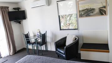 A hotel room with a bed, a desk with a chair, a TV, an air conditioner, a window with a view of trees, and a painting on the wall.