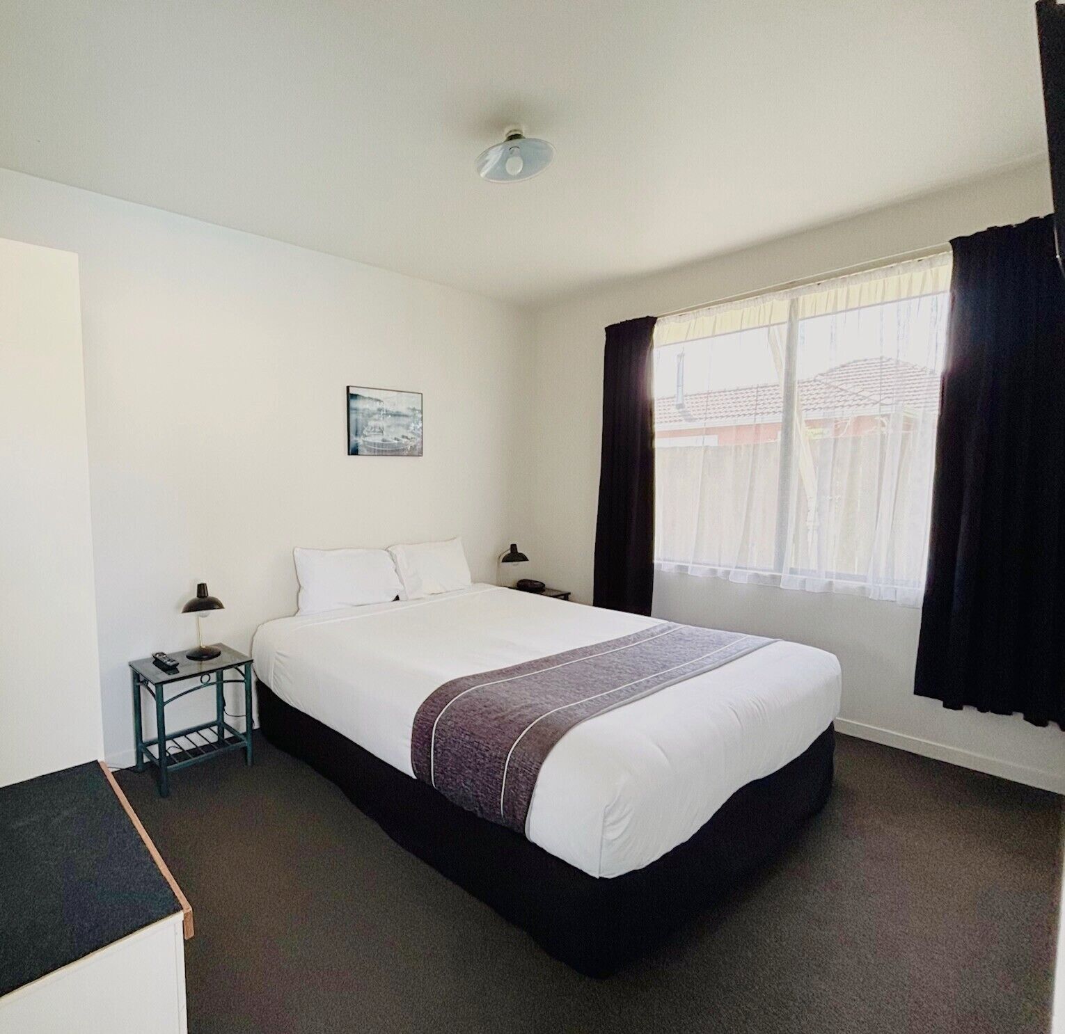 A hotel room with a bed, a small table with a lamp, a picture on the wall, and a window with curtains.