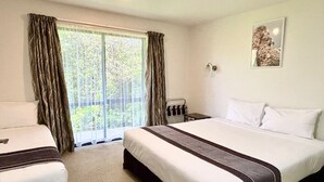 Habitación de hotel con una cama grande, dos camas más pequeñas, una ventana con cortinas, una mesita y una lámpara fijada a la pared.