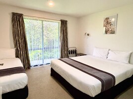 Habitación de hotel con una cama grande, dos camas más pequeñas, una ventana con cortinas, una mesita y una lámpara fijada a la pared.