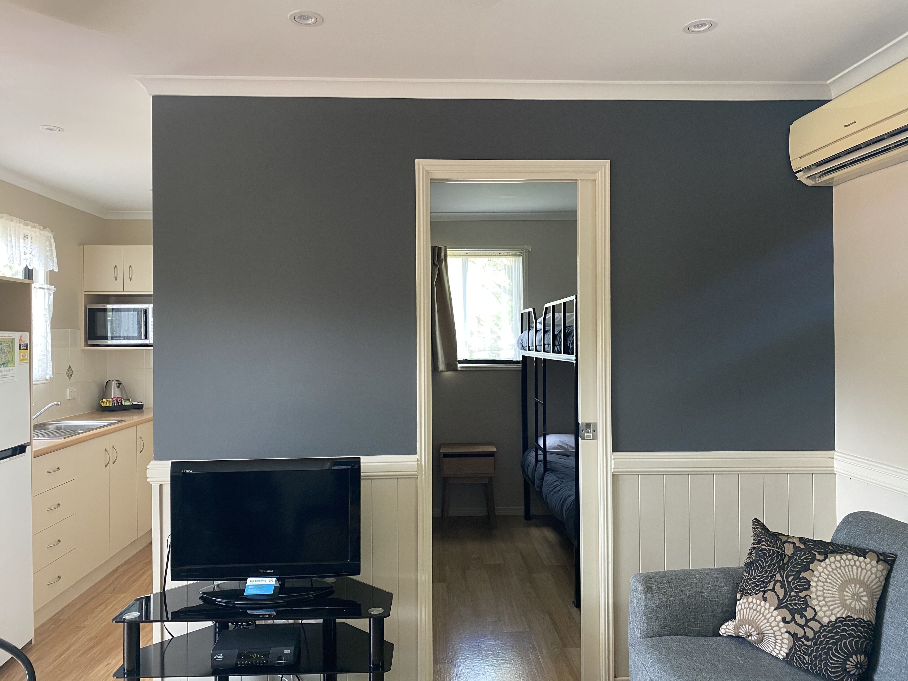 A compact living space with a kitchenette, a television on a stand, a grey sofa with a patterned pillow, and a dark wall.