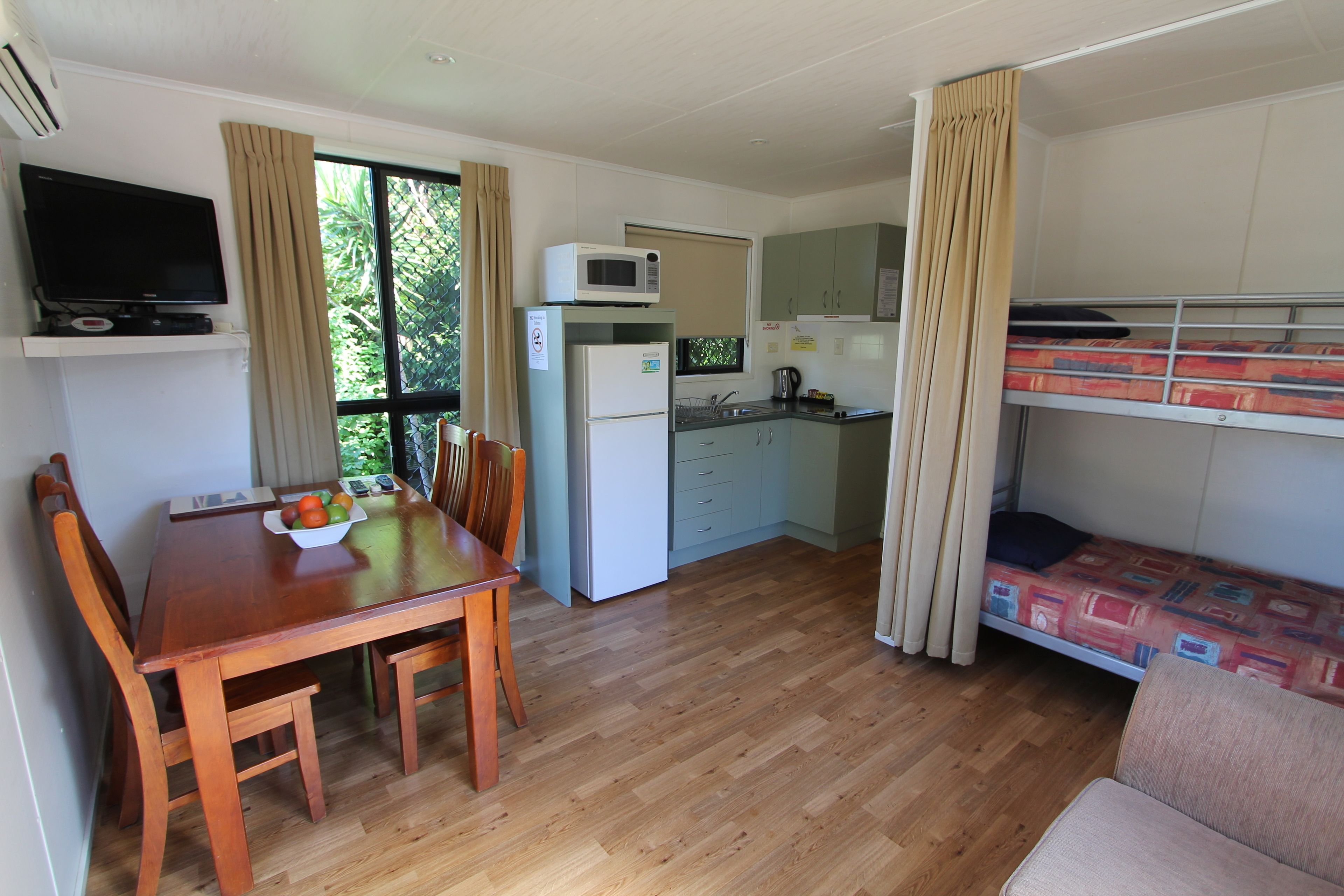 Een compacte kamer met een houten eettafel, stoelen, een kleine kitchenette met een magnetron, koelkast en wastafel. Er staat een stapelbed met een gestreept matras en een televisie aan de muur bevestigd.