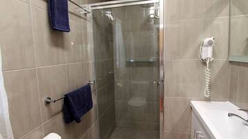A bathroom with a glass shower enclosure, a white sink, a hairdryer, and two towels hanging on hooks.