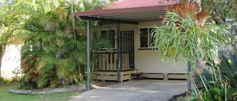 A small, single-story house with a porch, surrounded by lush tropical vegetation.