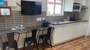 A modern kitchen with a flat-screen TV, a microwave, and a kettle on the counter. There are three black chairs and a small table.