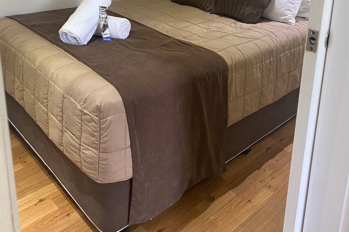 Una cama individual con un edredón marrón y almohadas blancas, una toalla blanca enrollada y un folleto pequeño sobre la cama.