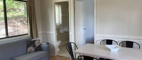 Una habitación con un sofá gris, una mesa de comedor con cuatro sillas, un ventilador de techo y vistas a un jardín a través de una ventana.